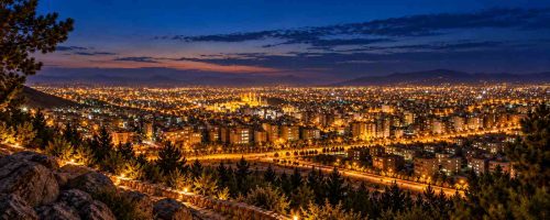 نمای پانوراما از بام مشهد در شب با چراغهای روشن شهر، ساختمانها و چشمانداز کوهستانی پارک خورشید